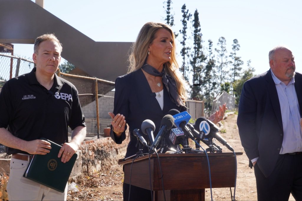 Administrator Loeffler alongside with other government officials standing behind a podium with microphones to address disaster survivors